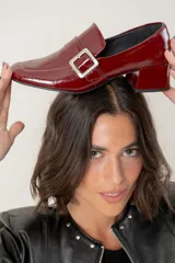Mocasin de cuero charolado color bordó, con punta cuadrada, taco bloque bajo y detalle de tira con hebilla metálica plateada en el empeine.