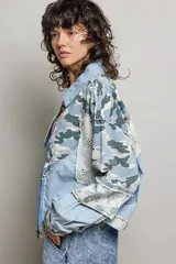 Chaqueta de denim de corte holgado y manga larga, con diseño en técnica de patchwork que combina sectores de mezclilla clásica, estampa de camuflaje, parches florales y detalles de encaje blanco. Cuenta con cierre frontal de cremallera metálica y cuello camisero.