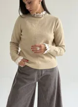 Sweater de punto color beige, con cuello redondo adornado con apliques de pedrería y detalle de puntilla blanca en el cuello y los puños.