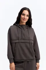 Hoodie de algodón color marrón con capucha, cierre frontal parcial y detalle de costuras horizontales en contraste negro. Presenta diseño holgado, mangas largas con puños elásticos y cordones ajustables en la capucha.