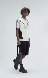 Chomba tejida de punto cerrado color off-white, con cuello polo y ribetes negros. Presenta un diseño deportivo con paneles laterales negros, número "90" y texto "Ravers" bordado en el frente.