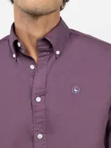 Camisa de hombre color azul, con cuello buttondown, cierre frontal con botones y logo bordado en el pecho. Presenta un acabado vintage logrado mediante el proceso de teñido en prenda.