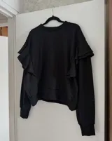 Sweater negro de textura acanalada con diseño de volados en los hombros y mangas largas. Presenta un corte holgado y cuello redondo.