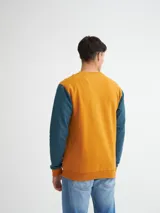 Buzo azul con cuello redondo, mangas largas y puños y bajo acanalados. Presenta bloque de color naranja en hombros y laterales, y estampado frontal con texto en color borgoña.