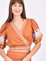 Blusa color ladrillo con escote cruzado, mangas cortas abullonadas con detalle de estampado de ondas en lila y naranja, y ribete de conchillas en escote, cintura y mangas.