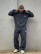 Conjunto de dos piezas compuesto por un buzo con capucha y cierre frontal y un pantalón de jogging, ambos confeccionados en tejido con efecto acid wash en color gris oscuro. El buzo cuenta con bolsillos frontales y logo bordado, mientras que el pantalón presenta un corte holgado y logo bordado en la pierna.