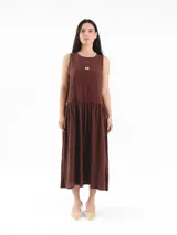 Vestido largo sin mangas de color marrón, de calce amplio y relajado, con falda fruncida y escote redondo. Cuenta con detalles de bordado a contratono en las sisas y logo frontal.