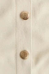 Cárdigan corto de punto fino color crema, con cuello redondo, cierre frontal con botones dorados y bolsillos delanteros decorativos con botón.