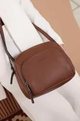 Bandolera de diseño compacto en color beige, confeccionada en material símil cuero con textura granulada. Cuenta con un bolsillo frontal con cierre y correa larga regulable.