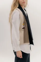 Chaleco abierto de tejido de punto, con diseño de bloques de color verticales en gris claro, negro y gris oscuro. Presenta un cuello ancho tipo chal y un detalle de tres pequeñas aplicaciones bordadas horizontales en tonos verde oliva, naranja quemado y celeste pálido en el frente.