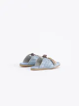 Zapatilla tipo mule (sin talón) de cuero vacuno, color celeste con efecto gastado y detalles de recortes. Presenta un logo estilizado en el lateral y cordones anchos de color beige y marrón.