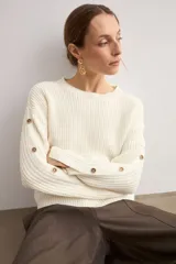 Sweater de punto color blanco natural, con cuello redondo y mangas largas adornadas con botones dorados a lo largo de las costuras exteriores.