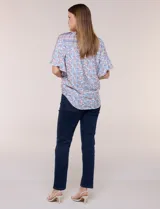 Blusa holgada de poliéster con estampado floral y geométrico en tonos celestes, azules y anaranjados sobre base blanca. Presenta cuello a la base con escote en V y botones frontales, mangas cortas amplias de estilo mariposa y un nudo en el ruedo.