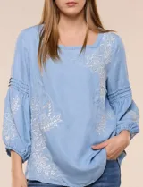Blusa de tencel celeste con escote cuadrado, mangas tres cuartos abullonadas con puños elastizados y delicados bordados florales blancos en el frente y las mangas.