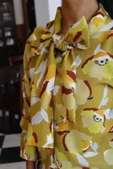 Blusa de manga larga con estampado floral en tonos mostaza, amarillo, blanco y bordó. Presenta un cuello con lazo integrado para anudar al frente.