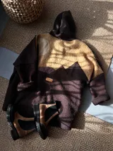 Campera con capucha de diseño patchwork, confeccionada en tejido tipo corderito en tonos marrón y tostado. Presenta un diseño que simula un paisaje montañoso con cortes irregulares y etiqueta de marca en la parte posterior.