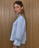 Blusa blanca de algodón con escote en V y cuello mao. Presenta mangas largas abullonadas con puños acampanados y fruncidos.