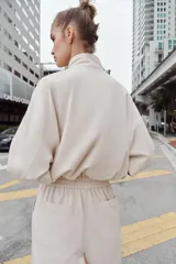 Cazadora estilo bomber de color beige, con cuello subido, manga larga y cierre frontal con cremallera. Presenta bolsillos delanteros de vivo y bajo ajustable con cordones elásticos laterales.
