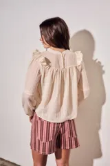 Blusa de color arena con diseño romántico, presenta volados en los hombros, detalles de encaje en el pecho y mangas largas con puños elásticos.