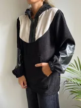 Canguro de pana negro y blanco con capucha, cierre frontal parcial con cremallera y mangas largas con puños elásticos. Presenta un bolsillo tipo canguro en la parte delantera y detalles en cuero sintético negro en la cremallera, la capucha y los codos.