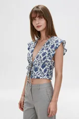 Blusa de diseño estampado con motivos florales y cachemira en tonos marrones sobre base blanca, con escote en V profundo y doble lazo frontal ajustable. Presenta mangas cortas con volados.