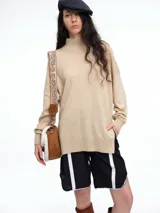 Sweater de tejido de punto color beige, con cuello alto, corte amplio y aberturas laterales en el ruedo. Presenta un diseño de costura central vertical en el frente y mangas largas con puños acanalados.