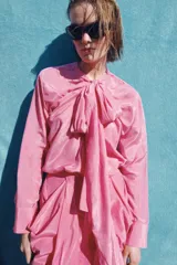 Blusa fluida de color rosado con cuello redondo ajustable mediante lazadas del mismo tejido, manga larga con puño y bajo asimétrico con aberturas laterales.