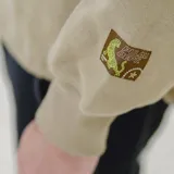 Canguro color marrón con capucha, cierre frontal parcial, bolsillo canguro y cordón ajustable en el dobladillo. Tiene un estampado blanco en el pecho con el texto "PULAU DESIGN DEPT." y un parche amarillo en la manga izquierda.