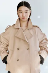 Gabardina corta color beige, de estilo cruzado con doble botonadura, solapas de muesca, bolsillos ribeteados y mangas raglán con charreteras en los hombros.