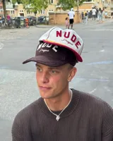 Gorra de béisbol de cinco paneles color blanca con visera color bordó. Presenta un parche frontal de fieltro con la inscripción "NUDE Project" y bordados de hojas en la visera en color amarillo mostaza.