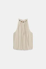 Top de cuello halter color beige, con detalle de volantes y abalorios de diferentes colores en el cuello. El bajo está acabado en elástico y tiene cierre en la espalda con abertura y lazada.