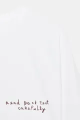 Camiseta blanca de manga larga con cuello redondo y texto bordado en color rojo en el pecho.