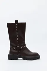Bota de caña media estilo biker, confeccionada en cuero sintético color marrón oscuro. Presenta una suela track robusta, detalle de tachas metálicas verticales a lo largo de la caña y un arnés decorativo con argolla metálica en el tobillo.
