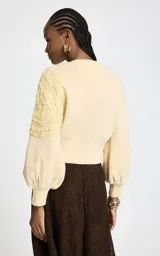 Cardigan de punto color amarillo claro, con cierre frontal de botones metálicos, cuello redondo y mangas largas abullonadas con puños ajustados. Presenta un diseño asimétrico con un panel de punto calado texturizado aplicado sobre uno de los hombros.