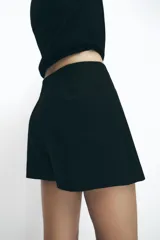 Falda pantalón negra de tiro alto con diseño cruzado frontal, dos botones dorados decorativos y falsos bolsillos de doble vivo.