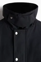 Campera negra de sarga encerada de algodón con cuello de pana. Presenta cierre frontal cubierto por una solapa con botones a presión, mangas raglán con puños ajustables mediante botones a presión, bolsillos insertados al bies y un bolsillo interior con botón a presión. Tiene un corte holgado y forro interior.