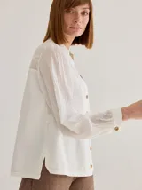 Camisa de corte recto y manga larga, confeccionada en algodón blanco. Presenta un canesú superior con bordado inglés floral y detalle de placa metálica con logo en la espalda.