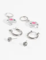 Set de aros plateados con tres pares diferentes: aros colgantes con dije ovalado con diseño de flor rosa, aros tipo argolla y aros tipo botón con strass.