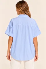 Camisa de algodón celeste, de corte recto, con cuello camisero, escote en V y un botón dorado decorativo en el centro. Cuenta con mangas cortas con vuelta y diseño holgado.