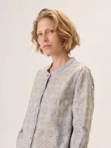 Camisa de algodón color gris claro, con cuello mao, cierre frontal de botones y mangas largas. Presenta un estampado sutil de hojas y flores, y detalles de bordado calado en los hombros.