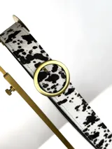 Cinturón de cuero genuino con estampado animal print tipo vaquita en blanco y negro. Presenta una hebilla circular metálica plateada.