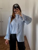 Camisa de rayas verticales celestes y blancas, con cuello alto y cierre frontal de botones. Presenta detalles de puntilla blanca en el cuello, a lo largo de la botonera y en los puños. Tiene mangas largas con terminación acampanada y diseño holgado.