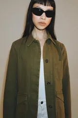 Chaqueta de estilo utilitario color verde militar, con cuello solapa, manga larga y cierre frontal con botones ocultos por solapa. Presenta bolsillos con solapa en la parte delantera y un bolsillo de plastrón en el pecho.