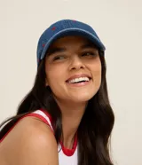 Gorra de denim azul con visera curva y pequeños corazones rojos bordados en toda la superficie. Cuenta con hebilla metálica ajustable en la parte posterior.