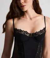 Corset de media copa en color blanco, confeccionado con paneles de poliamida y detalles decorativos de encaje floral en el borde superior y tul en los laterales. Cuenta con tirantes finos ajustables y varillas internas para estructura.