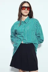 Camisa de silueta amplia y caída fluida, color celeste con rayas verticales blancas. Presenta cuello camisero clásico, cierre frontal con botones y un bolsillo aplicado en el pecho. Las costuras están resaltadas en azul oscuro.