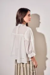 Blusa de algodón peinado color off-white, con cuello mao, cierre frontal de botones, mangas tres cuartos abullonadas con puños con volados, detalles de alforzas y apliques de encaje en el frente y los hombros.