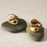 Anillo ajustable de acero inoxidable con baño de oro, con diseño abierto de doble gota. Una de las gotas presenta una incrustación de símil perla o piedra ojo de tigre, mientras que la otra es de metal liso pulido.