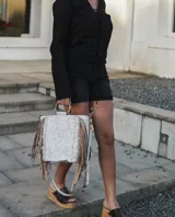 Cartera de mano tipo bolso con flecos laterales, confeccionada en cuero con acabado texturizado en tono claro. Presenta asas circulares metálicas, correa larga ajustable y extraíble, y un diseño de estilo boho.