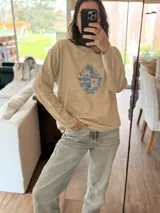 Sweater de punto color beige con cuello redondo y diseño de escudo bordado en el frente. Presenta un corte holgado y mangas largas.